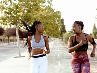 Two young fit black women running outdoors in a park in a sunny day while chatting. They wear sportswear and have braided hairstyle.