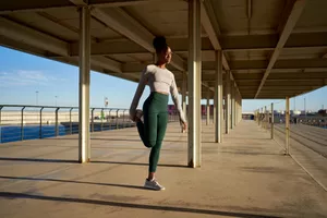 Full body of young female athlete with dark curly hair in activewear and sneakers sitting on embankment at seafront and stretching legs against cloudless blue sky