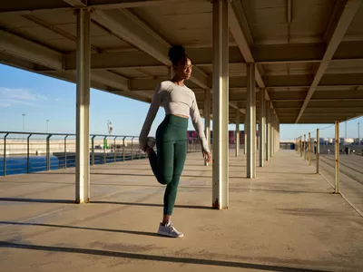 Full body of young female athlete with dark curly hair in activewear and sneakers sitting on embankment at seafront and stretching legs against cloudless blue sky