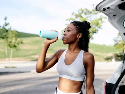 Attractive young black woman on sportswear and with braided hairstyle drinks water next to a car before she starts her training. She's outdoors next to a park in the daytime in a summer morning.