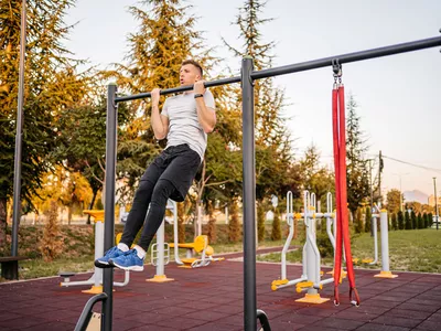 A handsome young man doing chin-ups on a bar outdoors in a public park.