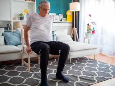 Senior man performing exercise inside his home with chair.