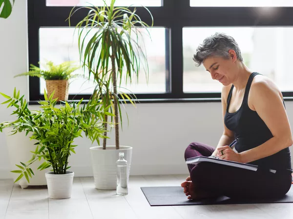 individual sitting on a yoga mat in fitness clothing near plants writing in a journal
