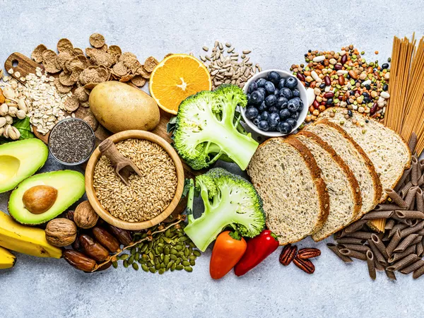 Overhead view of a large group of food with high content of dietary fiber arranged side by side. The composition includes berries, oranges, avocado, chia seeds, wholegrain bread, wholegrain pasta, whole wheat, potatoes, oat, corn, mixed beans, brazil nut, sunflower seeds, pumpkin seeds, broccoli, pistachio, banana among others. 