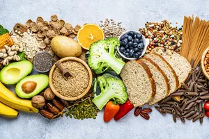 Overhead view of a large group of food with high content of dietary fiber arranged side by side. The composition includes berries, oranges, avocado, chia seeds, wholegrain bread, wholegrain pasta, whole wheat, potatoes, oat, corn, mixed beans, brazil nut, sunflower seeds, pumpkin seeds, broccoli, pistachio, banana among others. 