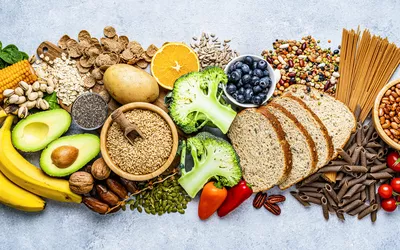 Overhead view of a large group of food with high content of dietary fiber arranged side by side. The composition includes berries, oranges, avocado, chia seeds, wholegrain bread, wholegrain pasta, whole wheat, potatoes, oat, corn, mixed beans, brazil nut, sunflower seeds, pumpkin seeds, broccoli, pistachio, banana among others. 