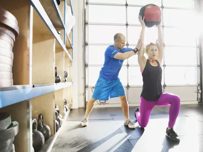 Gym instructor assisting woman with medicine ball lunge