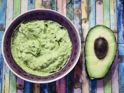 Bowl of guacamole and sliced avocado on colorful surface