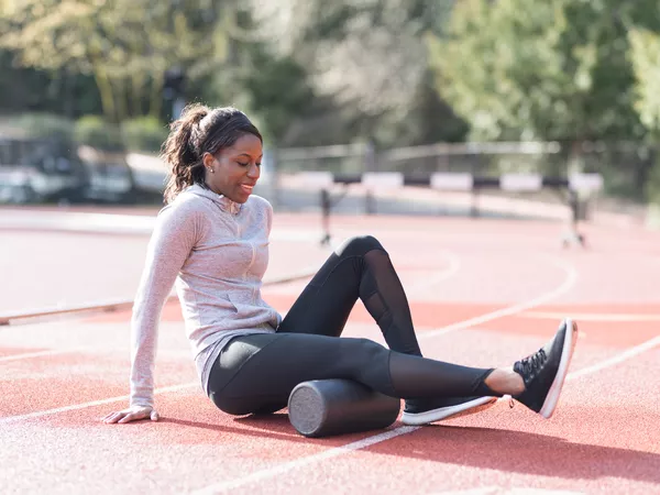 An African American athlete stretches out on the track before she starts her workout. She is sitting and has her legs out in front with a foam roller underneath the upper part of her leg.