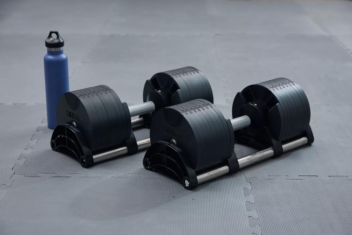 Nuobell Adjustable Dumbbells on stands next to water bottle on gray mat