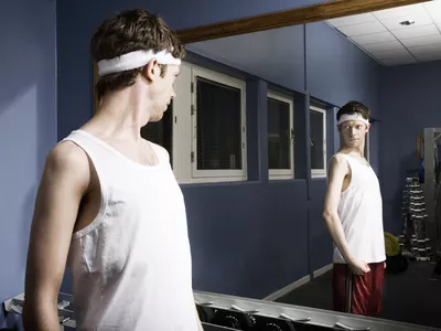 Man wearing sweatband looking at reflection in gym mirror, flexing arm