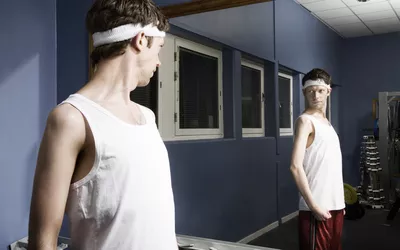 Man wearing sweatband looking at reflection in gym mirror, flexing arm