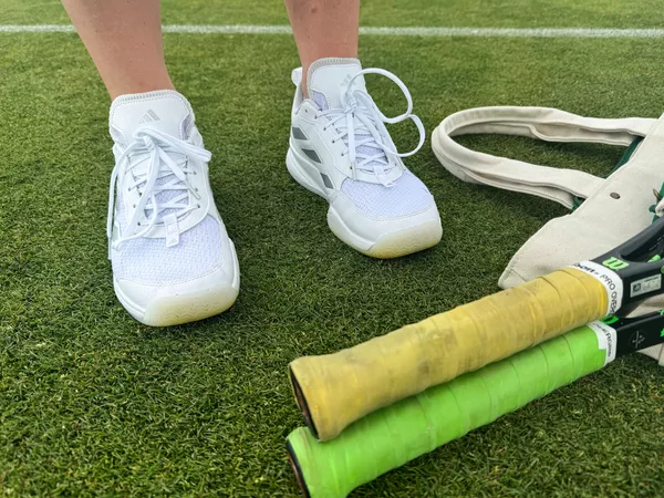 The Adidas Women's Avaflash Low Tennis Shoes worn on a grass court next to bag and rackets