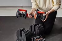 Hand holding FitRx SmartBells Set Quick Select Adjustable Dumbbells and kneeling in front of stand