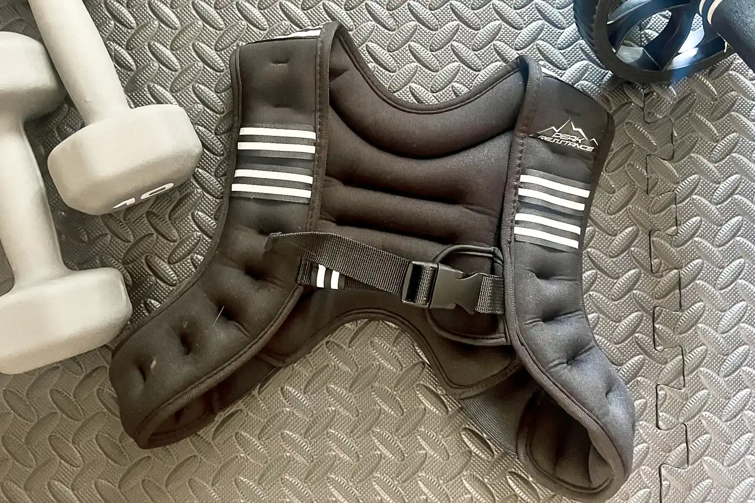 Aduro Sport Adjustable Weighted Vest placed on a textured mat surrounded by gym equipment including dumbbells and an ab wheel