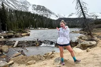 A person smiles and poses near a river wearing the Asics Women's Gel-Trabuco 13 Trail Running Sneakers