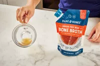 hand holds glass of Bare Bones Grass-Fed Beef Organic Bone Broth with pouch on countertop