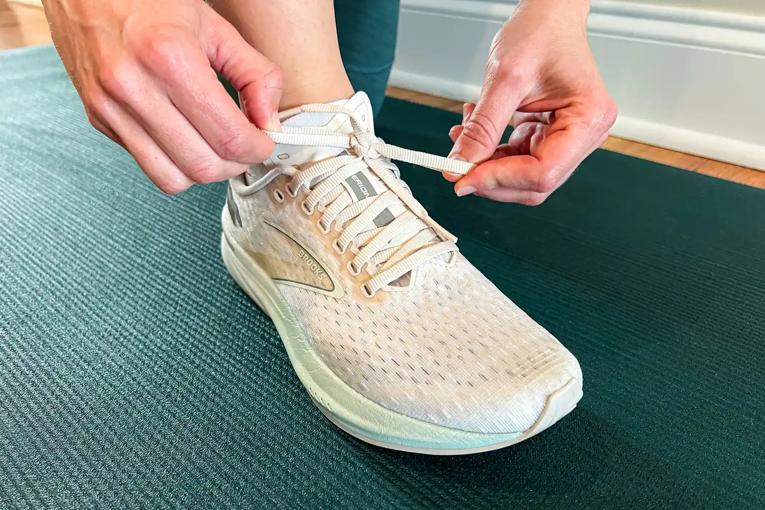 A person tying the laces of a running shoe labeled 'Brooks' on a yoga mat