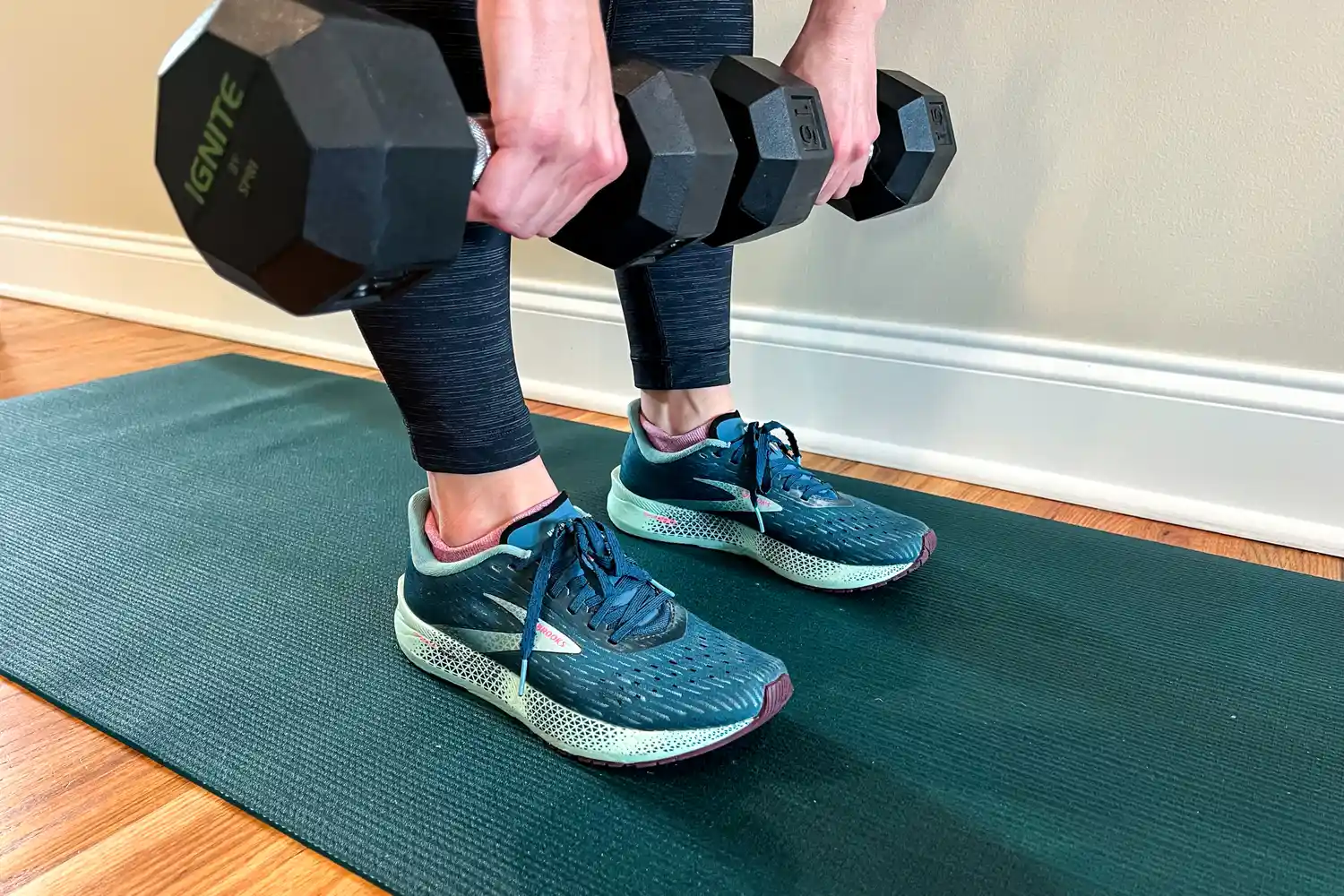 Close-up of hands holding dumbbells, green exercise mat and running shoes visible