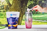 A hand adding a scoop of Gnarly Nutrition Hydrate Electrolyte Powder into water in a glass on an outdoor wooden table