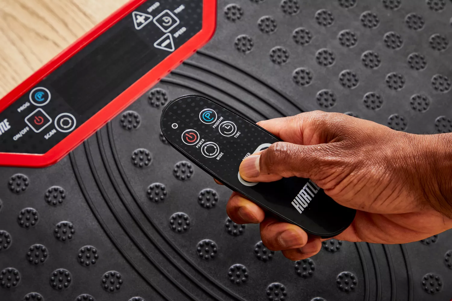 A hand uses a remote control to activate the Hurtle Fitness Vibration Platform Machine