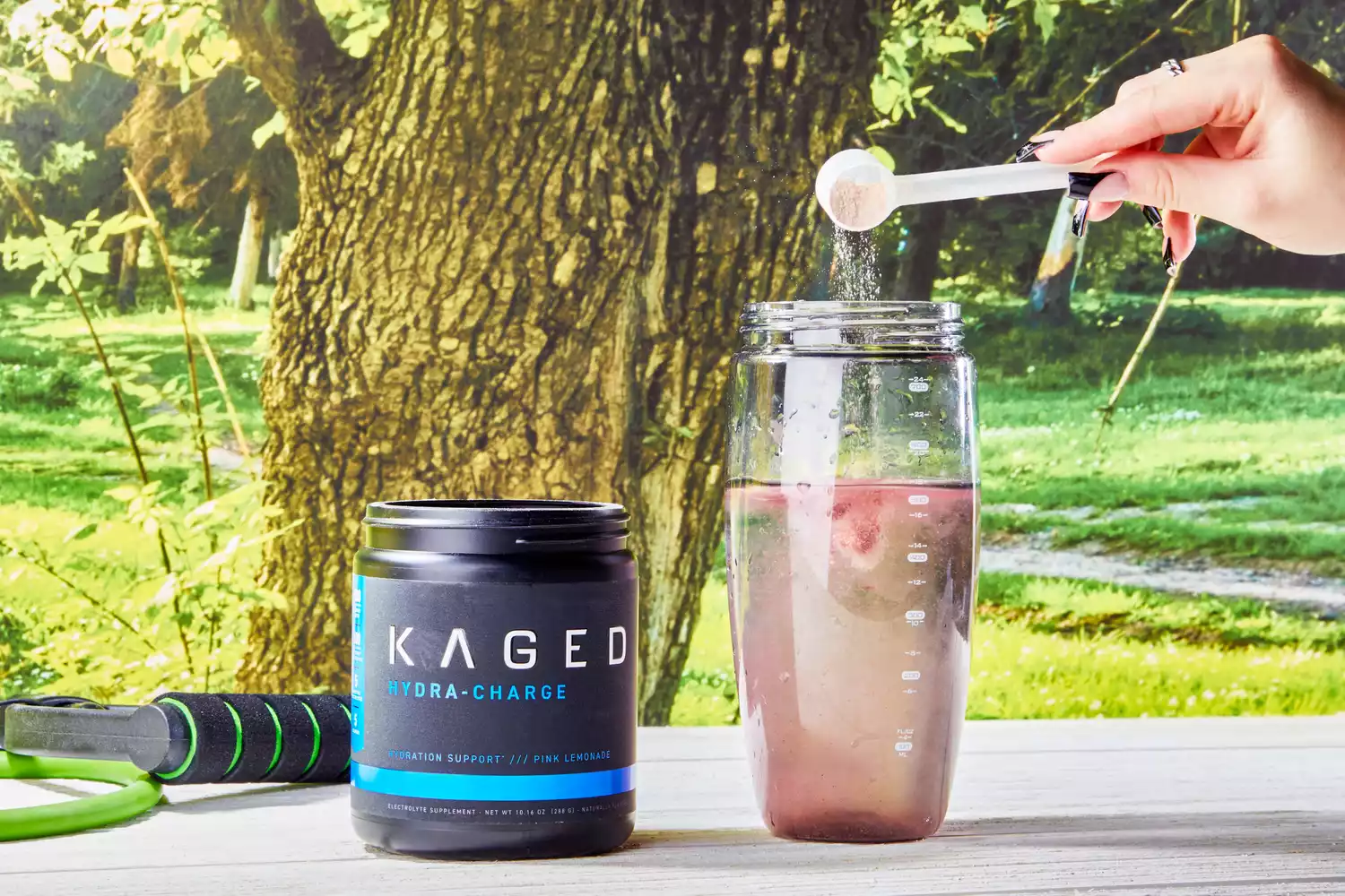 Kaged Hydra Charge jar next to a hand adding a scoop of powder into a blender bottle filled with water