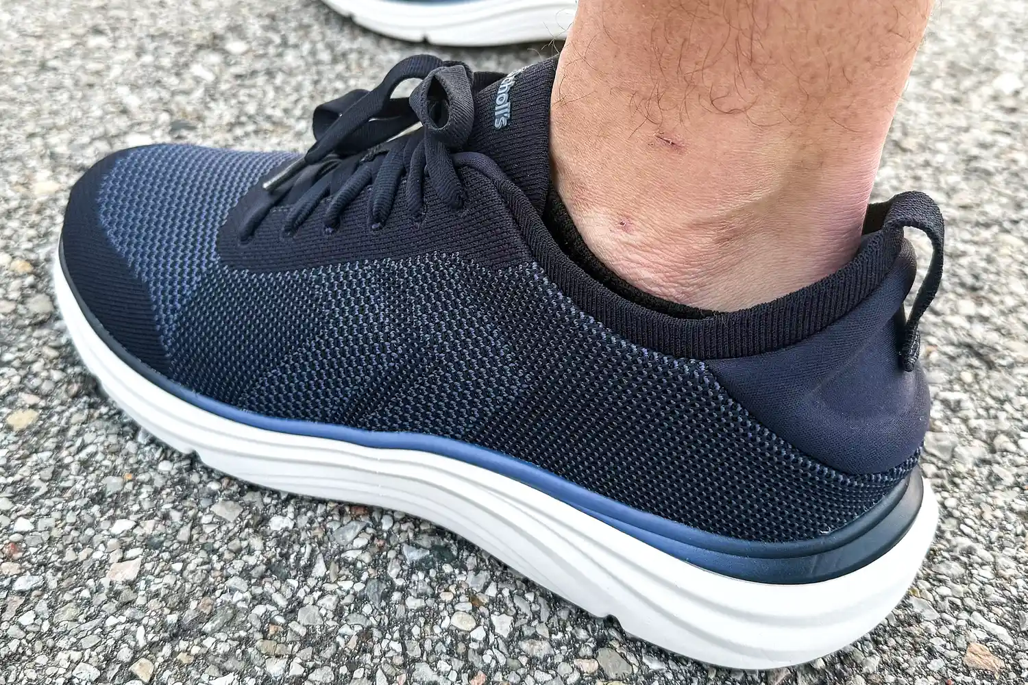 A foot wearing the Dr. Scholl's Men's Feel Ready Lace Up Sneakers on asphalt