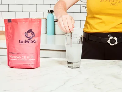 A hand using a spoon to mix Tailwind Nutrition Caffeinated Endurance Fuel into water in a glass