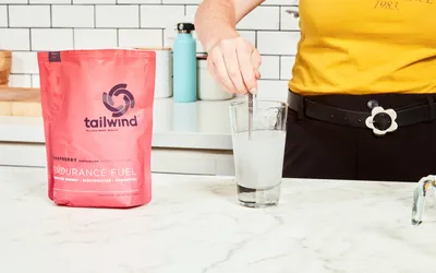 A hand using a spoon to mix Tailwind Nutrition Caffeinated Endurance Fuel into water in a glass