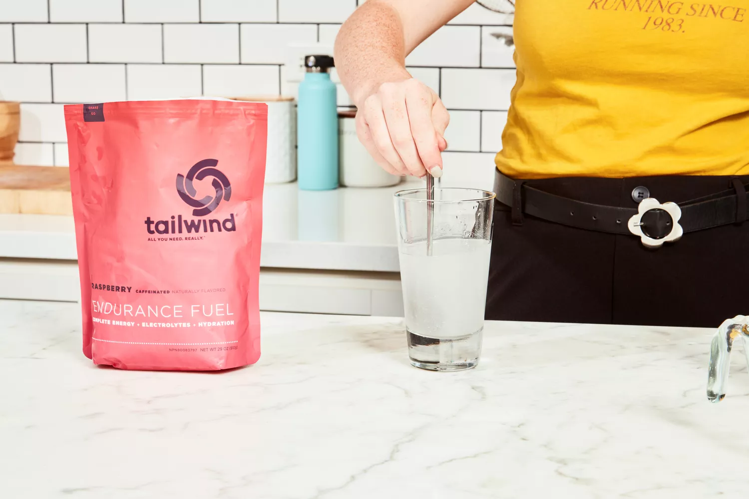 A hand using a spoon to mix Tailwind Nutrition Caffeinated Endurance Fuel into water in a glass