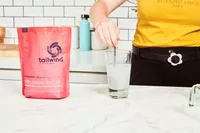 A hand using a spoon to mix Tailwind Nutrition Caffeinated Endurance Fuel into water in a glass