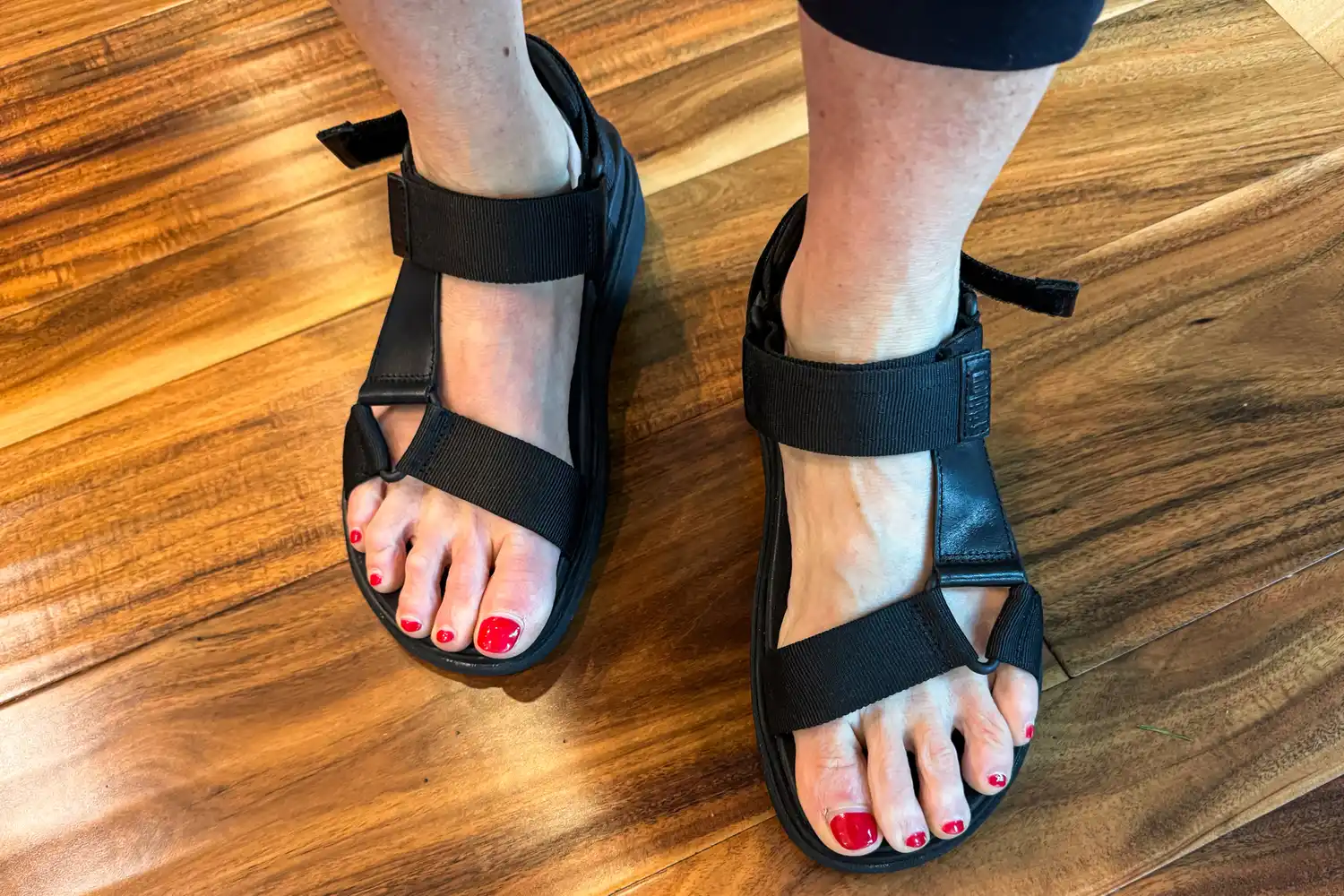 An overhead view of a person wearing FitFlop Women's GEN-FF Adjustable Webbing/Leather Back-Strap Sandals on a wooden floor