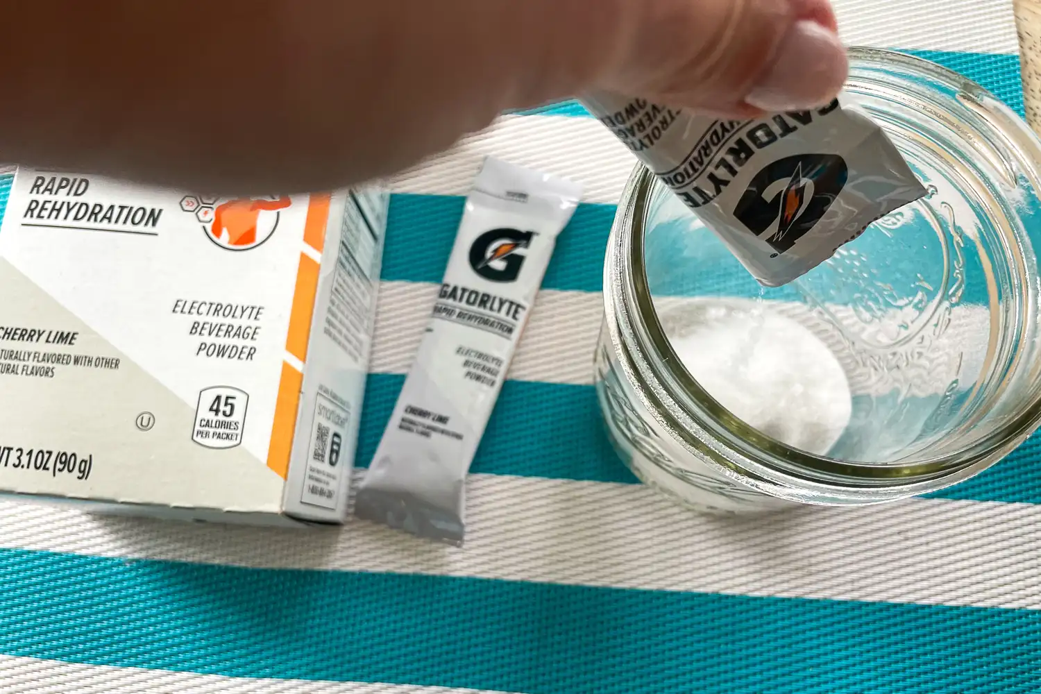 A hand pouring electrolyte beverage powder into a glass with packaging in the background