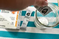 A hand pouring electrolyte beverage powder into a glass with packaging in the background