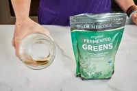 Person holding glass next to Dr. Mercola Fermented Greens Powder