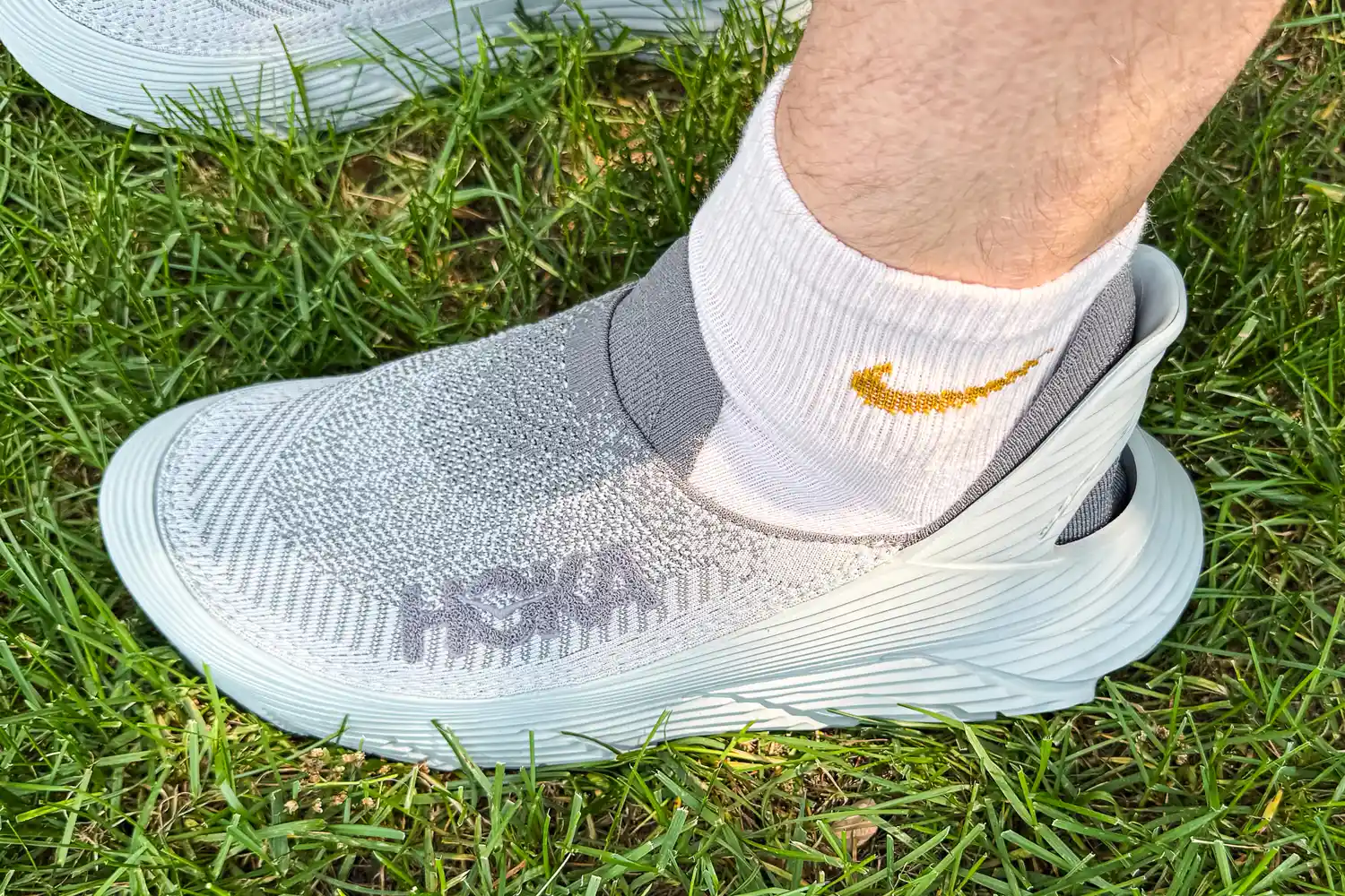 A person wears the Hoka Restore TC Recovery Shoes while standing in grass