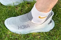 A person wears the Hoka Restore TC Recovery Shoes while standing in grass