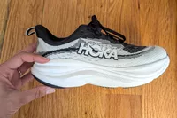 A hand holding a Hoka Women's Skyflow sneaker over a wooden surface