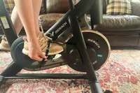 A person secures the pedals of the Merach S26 Exercise Bike to their sneakers