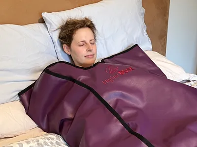 A person lying on a bed wrapped in an infrared sauna blanket with the Higher Dose logo visible