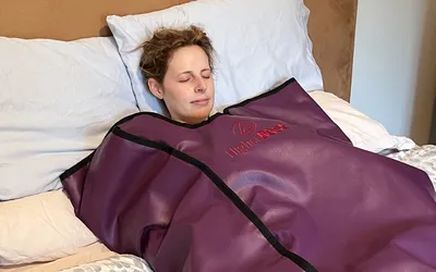 A person lying on a bed wrapped in an infrared sauna blanket with the Higher Dose logo visible
