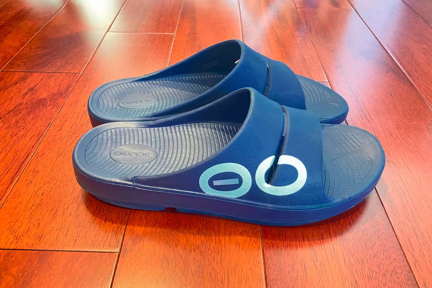 A pair of OOFOS OOahh Slide Sandals on a wooden floor