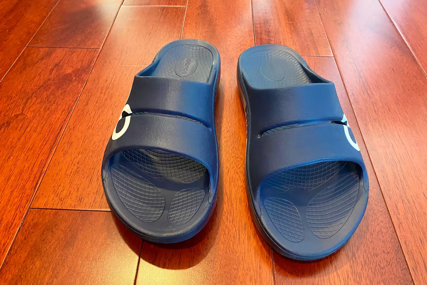 A pair of OOFOS OOahh Slide Sandals on a wooden floor