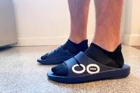 Feet wearing OOFOS OOahh Slide Sandals with socks on linoleum