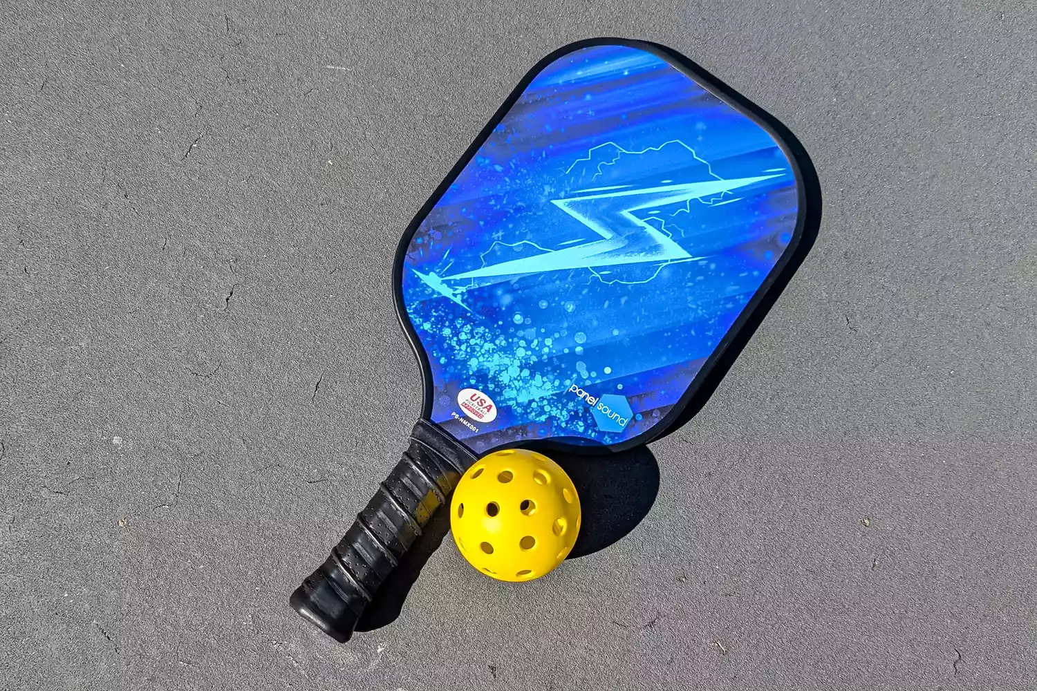 The Panel Sound Pickleball Paddle posed next to a ball on a gray surface