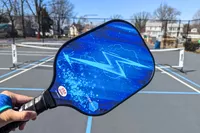 A hand holding a Panel Sound Pickleball Paddle on a court