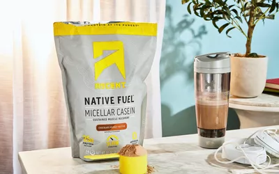 Ascent Native Fuel Chocolate Peanut Butter Micellar Casein Protein Powder in a scoop next to its container and blender bottle