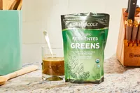 Dr. Mercola Fermented Greens Powder displayed on speckled counter in a glass next to its packaging