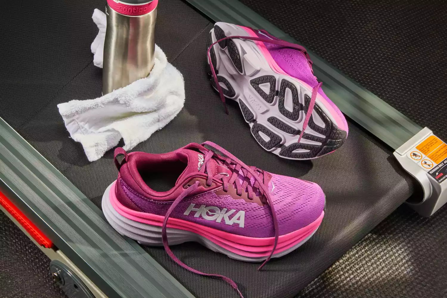 Pink Hoka Women's Bondi 8 Running Shoes displayed on a treadmill with a towel and water bottle