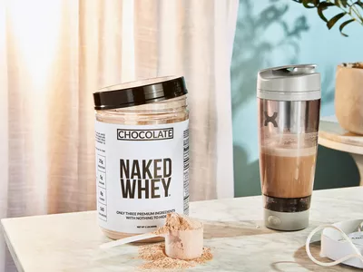 Naked Nutrition Chocolate Whey Protein Powder in a scoop next to its container and a blender bottle
